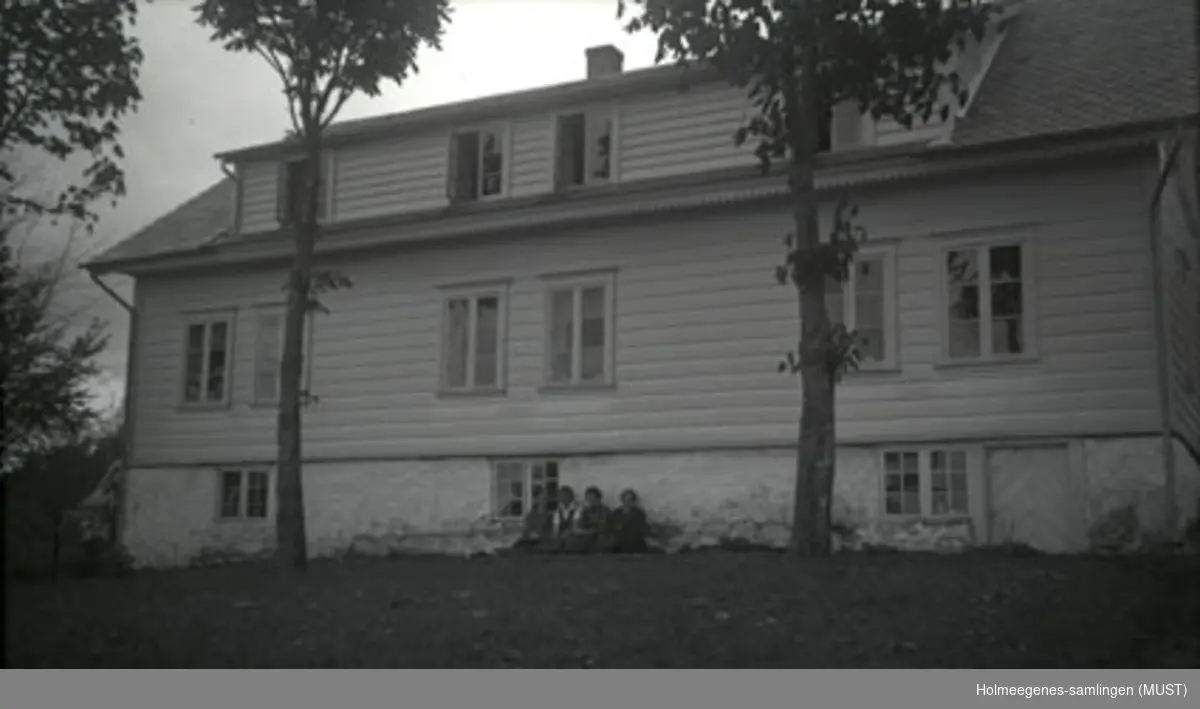 Et bolighus med kjeller og stor ark. Trær i forgrunnen. Fire unge kvinner sitter inntil husets grunnmur.
