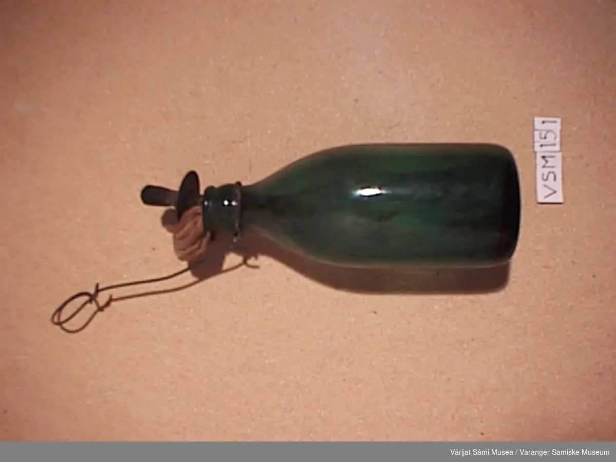Grønn glassflaske med veik av tøy. Veikholderen av metall og flaskeopphenget er av metall.