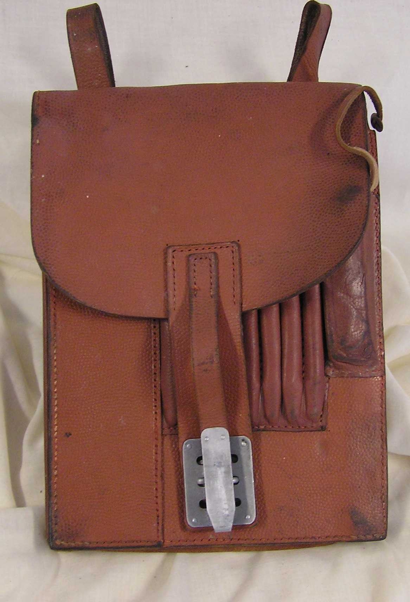 Teknikk: Kraftig, brunt lær. En innvendig lomme.Utvend.lomme inndelt i 7 smale rom f.blyanter(*)samt større,smal  *
Form: Rektangulær m/2 stropper øverst med metallspenner. En blyantrest i ene rom. Lomme for annet skriveutstyr.En klaff for luking. Denne har på- satt dobbelte lærstropper samt metallluking.  Utstyret har tilhørt de tyske tropper i Kragerø 1940-45.  
