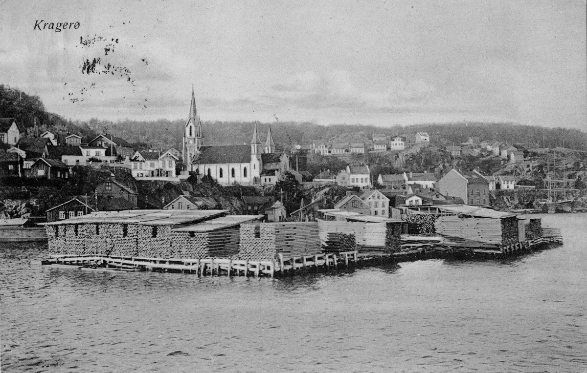 Postkort fra Kragerø. Trelast på brygga. Kirken i bakgrunnen