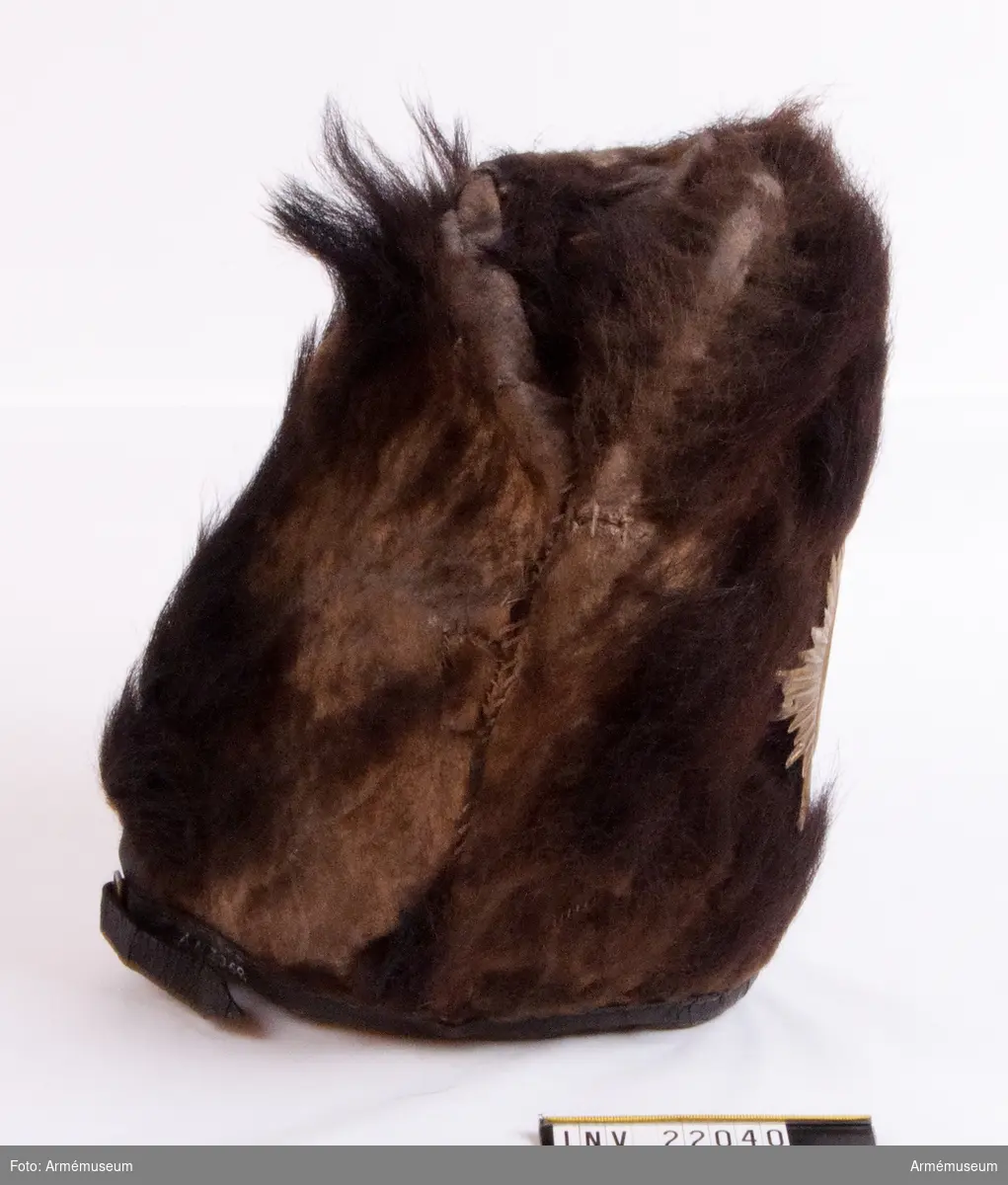Mössan, som är helt tillverkad av björnskinn, är stoppad på en spjälram med innerhatt av filt. 
Mössan saknar brätte men har en läderrem fastsydd i nederkant med ett mässingsspänne bak.