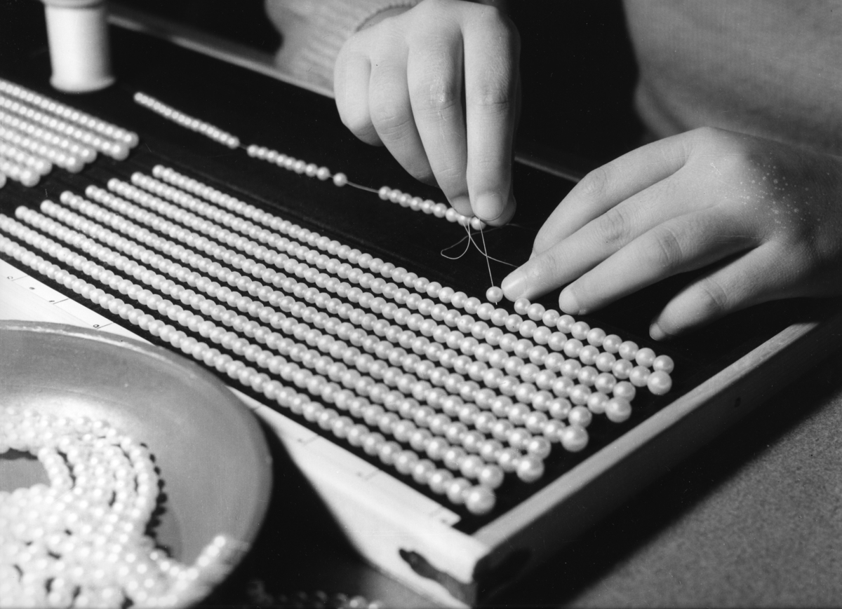 'Bilder till monter 254 med pärlhantering, i basutställningen.Tillverkning av pärlhalsband. ::  :: En person trär pärlor på nål och tråd.'