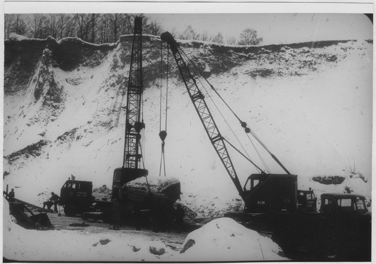 'Lastning av flyttblocket vid Dösebacka. Lastbilsförare Bo Andersson. Bildtext enligt Årstryck 1964: ''Fig. 1. Det vindslipade flyttblocket lyftes upp på vagnbjörnen. Grustagsväggen är ca 30 meter hög.'' Storleken på flyttblocket: Längd 7 m, bredd 5 m och höjd 2 m. Vikt ca 30 ton. :: Vindslipat flyttblock med ögongnejs, spår av glacialräffling och spår av vindslipning.'