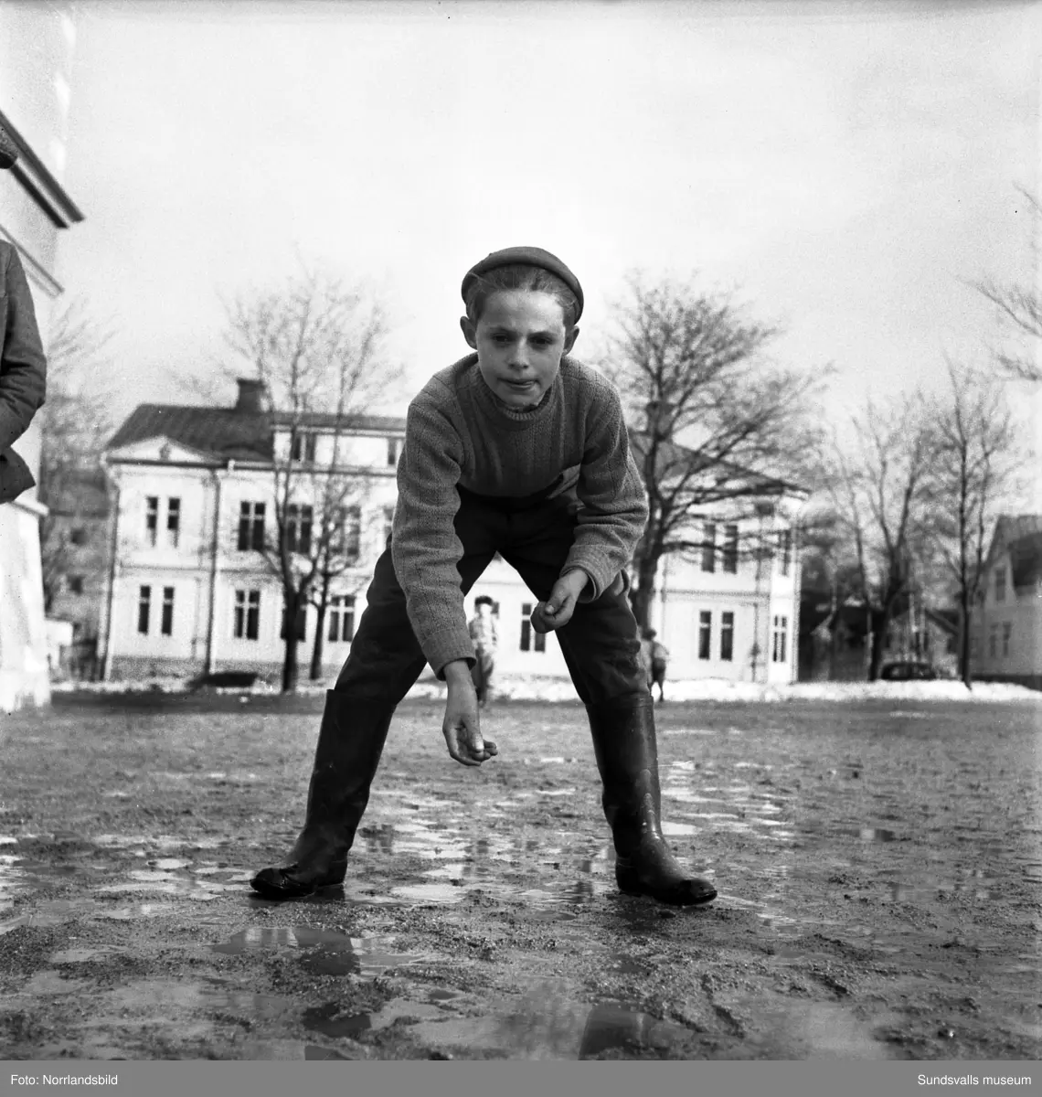 Flickor och pojkar på GA-skolan spelar kula, kastar boll och hoppar hage på en vårslaskig skolgård 1950. På en av bilderna bakom den kulspelande pojken syns det numera rivna huset på andra sidan Skolhusalln, kvarteret Borgaren 6. Nu (2015) är tomten obebyggd och används som parkering.
