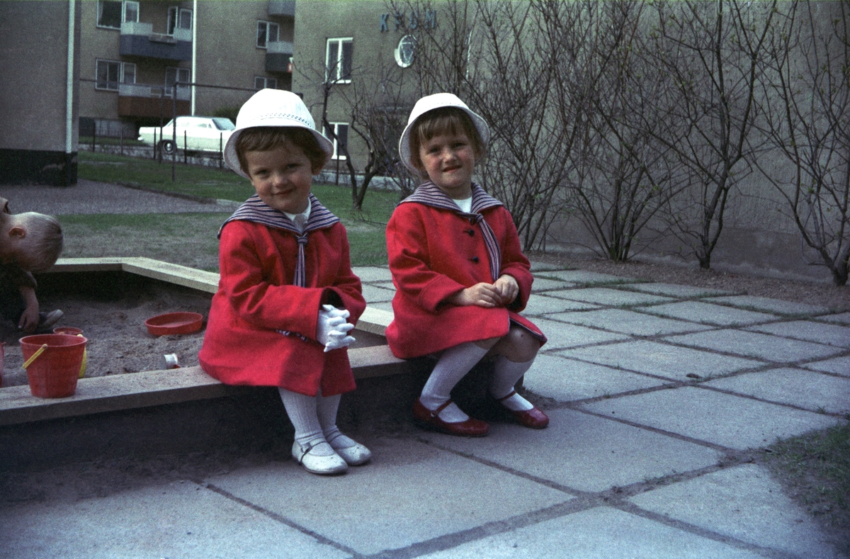Två små finklädda flickor sitter på kanten till sandlådan, som finns på gården vid bostadshusen.