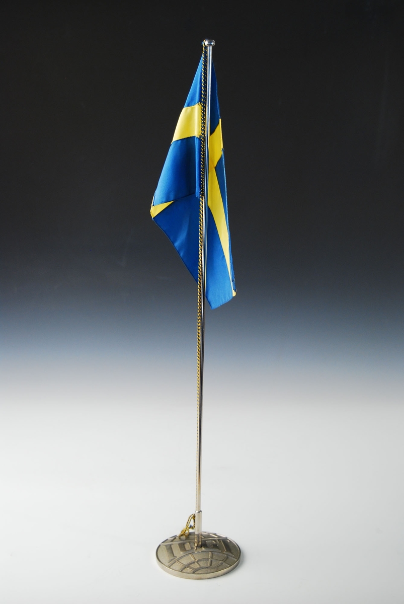 Svensk bordflagg. Sirkulær base som er utformet som IOGT (International Organisation of Good Templars) sitt symbol. Flaggstangen er festet i basen med ei skrue. Heisesnorene har blå og gul farge.