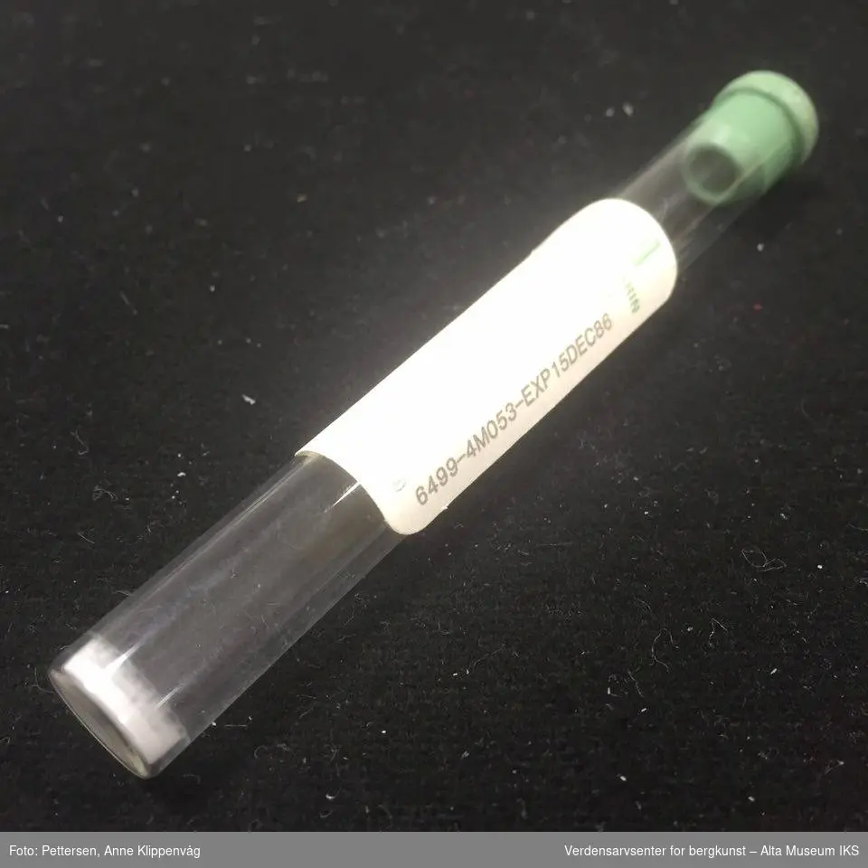 En sylindrisk beholder av glass, med grønn gummikork. I bunnen ser man et lite lag av hvitt pulver.
