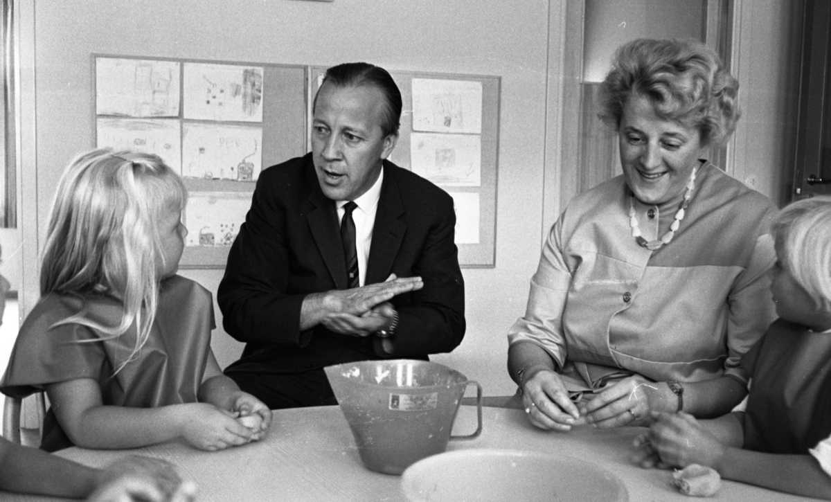 Barndaghem 9 september 1966

Vid ett bord sitter en man och en förskollärare med barn och leker med trolldeg. Bakom dem finns det teckninger på väggen.