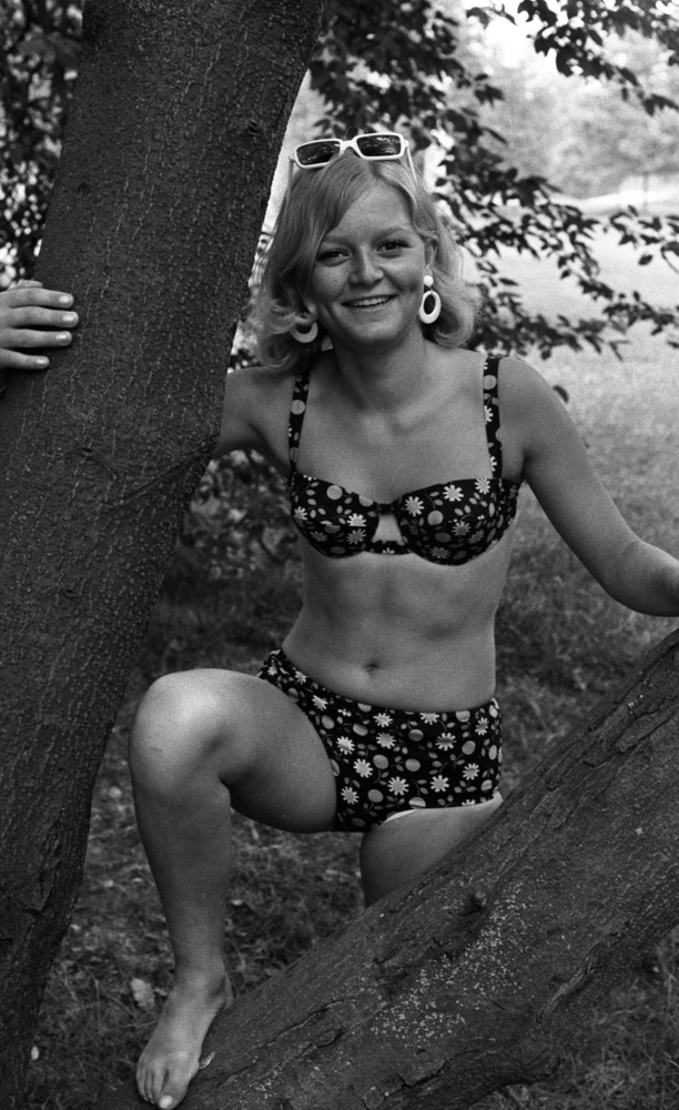 Baddräkter, Hus forts. 30 juni 1966

En fotomodell klädd i mörk blommig bikini står och poserar invid ett träd. Hon har ett par vita solglasögon uppe på huvudet samt ett par vita örhängen i form av stora clips med ringar.