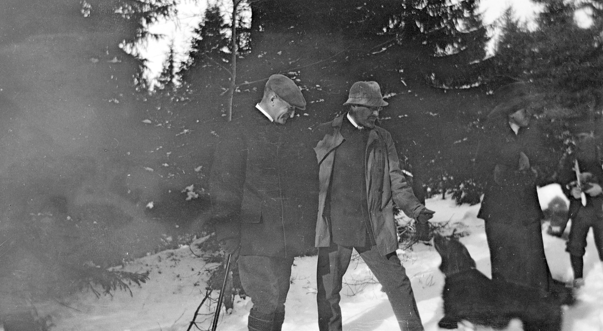 Et par mennesker står i snøen med en hund som hopper. Den ene av mennene  holder i en spaserstokk.