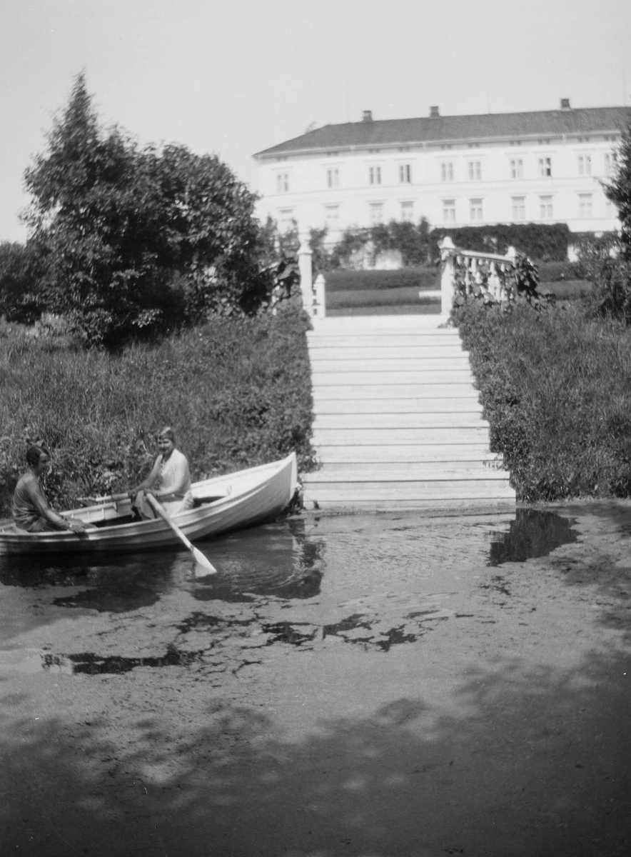 To kvinner i en robåt på en liten dam i hageanlegget ved sommerstid på Linderud Gård. I bakgrunnen sees hovedbygningen.