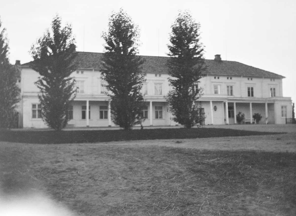 Forsiden av hovedbygningen på Linderud Gård. Foran huset står det beskårne trær i en rad.