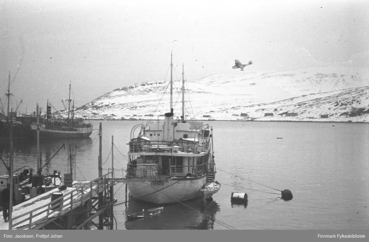 Hospitalskipet Elieser 4 utenfor Dampskipskaia i Hammerfest. En fraktebåt ligger ved kai og skorsteinen på det som sannsyligvis er Hurtigruta ses til venstre på bildet. Noen brakker er kommet opp på Fuglenes. De snødekte fjellene på bildet er Vardfjell til venstre og Storfjellet til høyre. Foten av Fuglenesfjell ses helt til høyre på bildet. Sjøen på bildet er blikkstille. I bakgrunnen lander et Catalinafly.