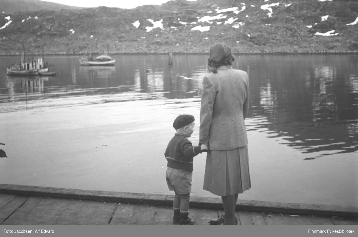 Arne Jacobsen og hans mor Aase fotografert på kaia i Havøysund. Aase har et ganske lyst skjørt og jakke på seg. Litt mørkere strømpebukse og mørke sko. På hodet har hun en lue eller tørkle. Arne har en mørk alpelue på hodet. Han har også en ganske mørk genser med en lys stripe rundt livet og håndleddene og lys shorts. På beina har han mørke sko og en ganske lys strømpebukse. Kaia de står på er av tre og en tverrplanke er slått på som et lite rekkverk. Tre fiskebåter ligger fortøy på den blikkstille havna. To av båtene ligger side om side helt til venstre på bildet. En liten, åpen trebåt er fortøyd på svai bak dem. Midt på bildet, over hodet på Arne, ses noen stolper ute på havna. Det er muligens en støtte/sjømerke. På andre siden av havna er et fjellparti. Terrenget består stort sett av stein og noen snøflekker ligger spredt rundt.