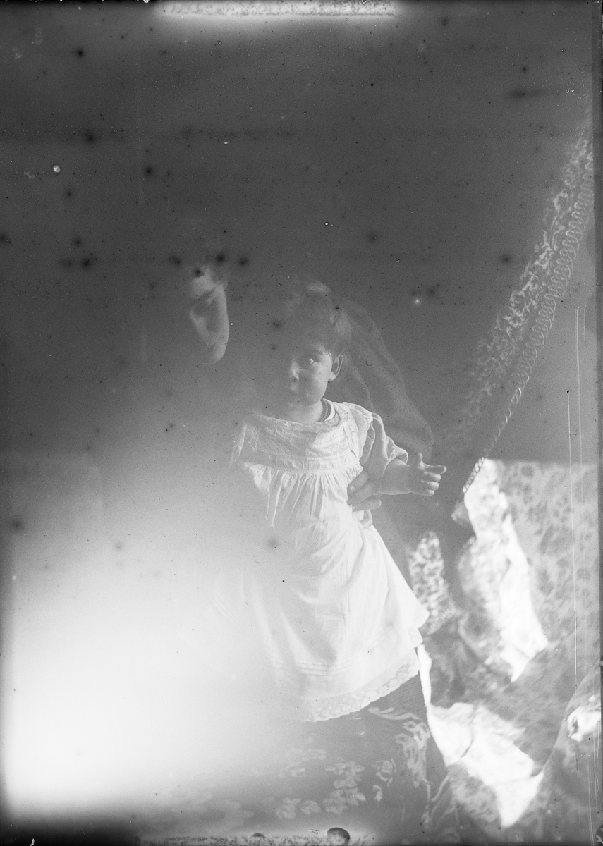 En kvinne holder et lite barn som står på en sofa. Lyset fra et vindu lyser opp motivet.