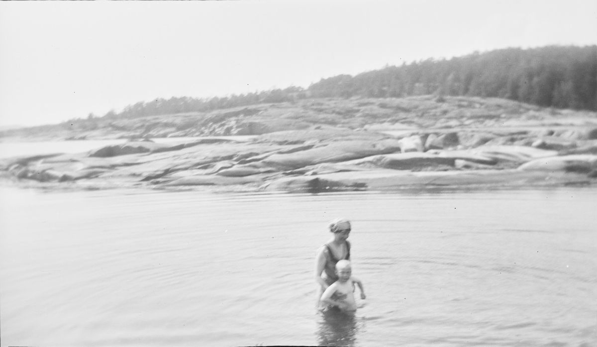 En kvinne står i vannet og holder en gutt under armene, som hjelp til å lære å svømme rygg. I bakgrunnen svaberg og skog.