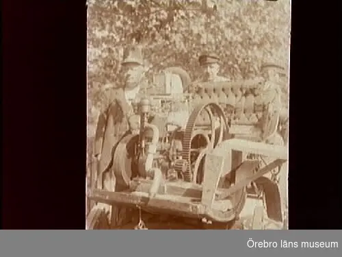 Två män och en maskin. 
Bilden är tagen vid Gate smedja. Mannen till vänster är David Johansson från Skrinhult.
Fordonet på denna bild är en hemmabyggd "automobil". Den är byggd i Gate, söder om Hjo, av smedmästaren Gustav Lindman och indelte soldaten Oscar Berg. Den är gjord av en gammal hästvagn och en encylindrig fotogenmotor, och sattes samman under natten till Kristi himmelsfärdsdag 1914. Den byggdes mest på skoj för att delta i ett "rally", men särskilt långt gick den dock inte. Den tog sig ett par kilometer från Gate smedja till Gate Handel, men sedan var sagan all.