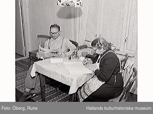 Maria och Knut Öberg sitter vid köksbordet. Maria skriver brev till sin bror i USA medan Knut läser tidningen. Bild 2: Porträtt av Maria när hon skriver brev.