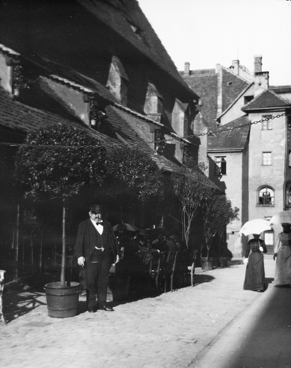Skioptikonbild med motiv man framför café på gata i Nürnberg. Bredvid promenerar två kvinnor med paraply.
Bilden har förvarats i kartong märkt: Nürnberg VI. 1901