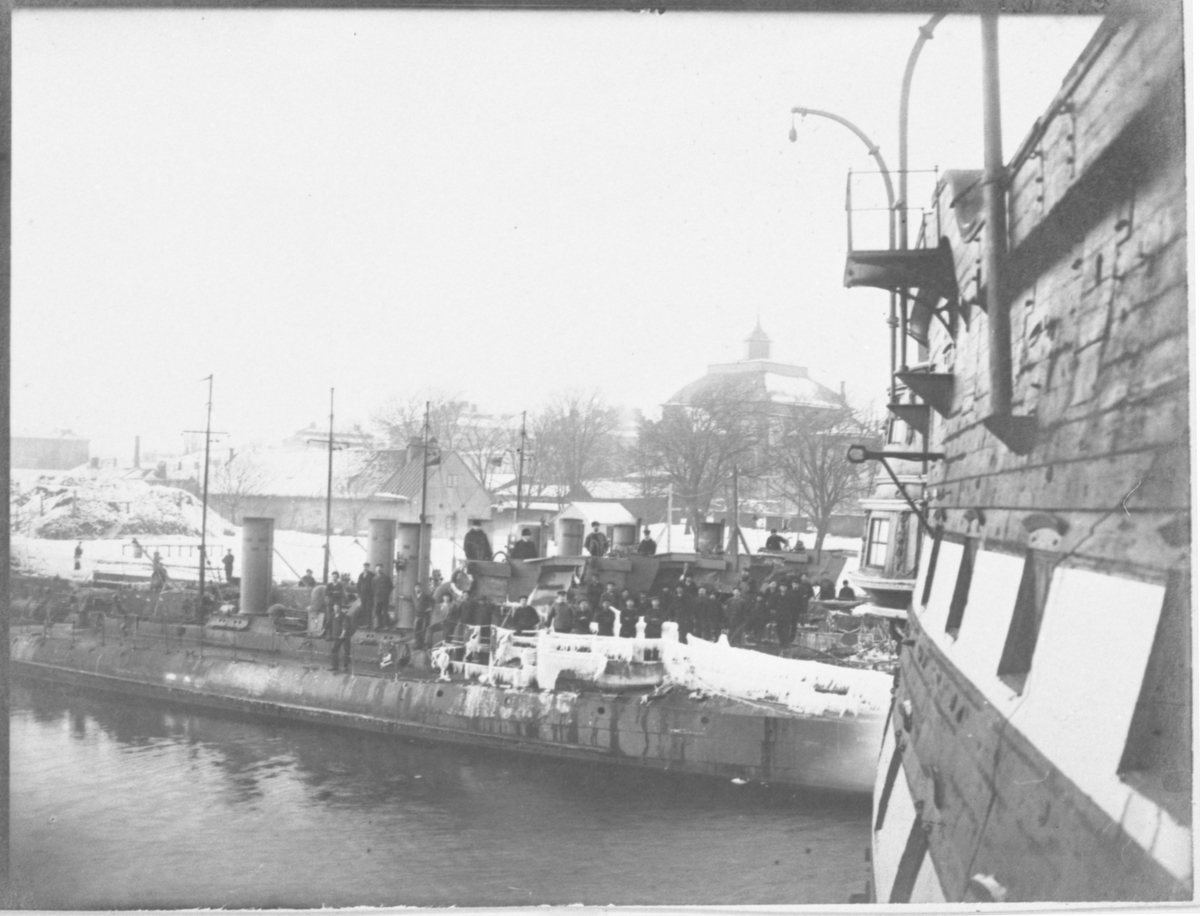 Bilden visar torpedbåtar på vinter som ligger förtöjt vid Kungsbron i Karlskrona. På däck står soldater. Åt höger sidan syns bordväggen av linjeskepp Stockholm som logementsfartyg. I bakgrunden ser man Amiralitetskyrkan.