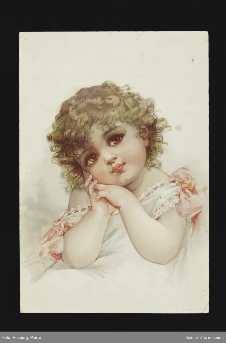 En liten flicka, sitter möjligen i en säng. Håller huvudet på sned, lockigt hår, klädd i vita kläder med rosa detaljer och rosetter.