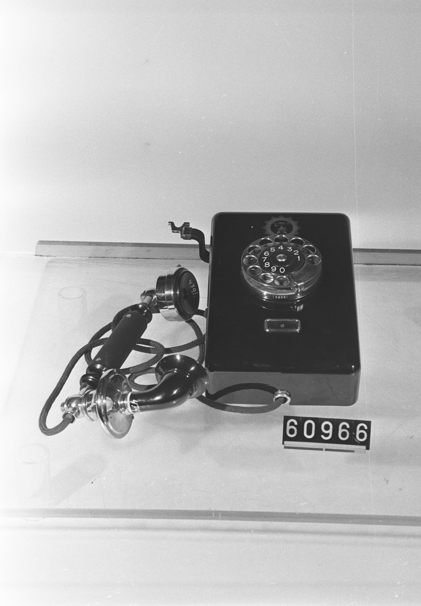 Telefonapparat DE 110 för AT-system. Väggmodell av svartlackerad järnplåt, 1922 års modell, med hängande mikrotelefon typ RE 4301, textilklätt mikrotelefonsnöre, fingerskiva av förnicklad mässing och apparatklocka med likströmsresistans 1000 ohm.