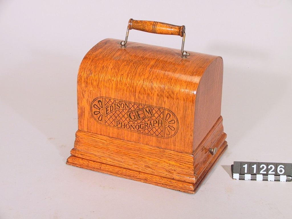 Fonograf, enligt Edison. Mindre typ, fjäderverksdriven, jämte "recorder" för upptagning. Recorderns nr. 10882. Monterad på träplatta. Skyddshuv med handtag.