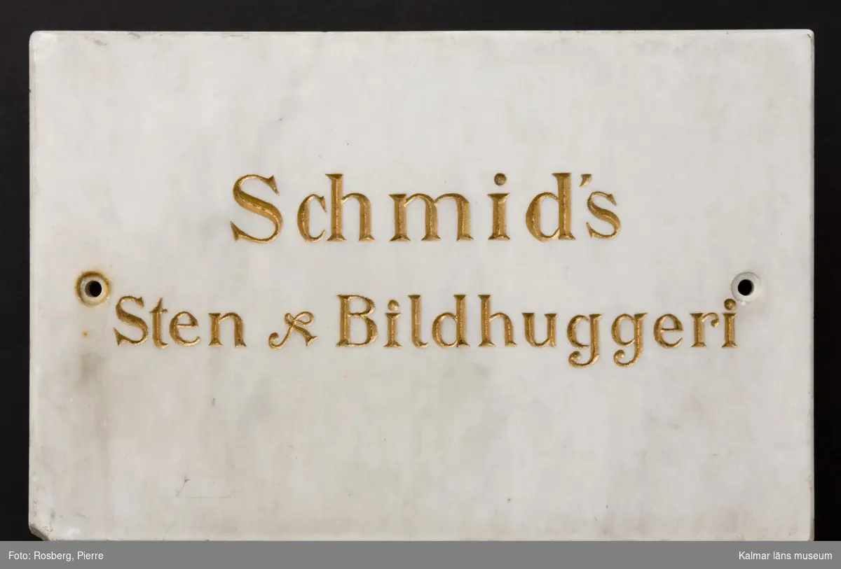KLM 12433. Skylt, stenhuggarskylt, bildhuggarskylt. Av marmor med text i guld. Marmortavla med försänkta bokstäver i guld. Text på båda sidor, på ena sidan: J.A. Schmid, Bildhuggare en Tr. upp, på andra sidan: Schmid´s Sten & Bildhuggeri.