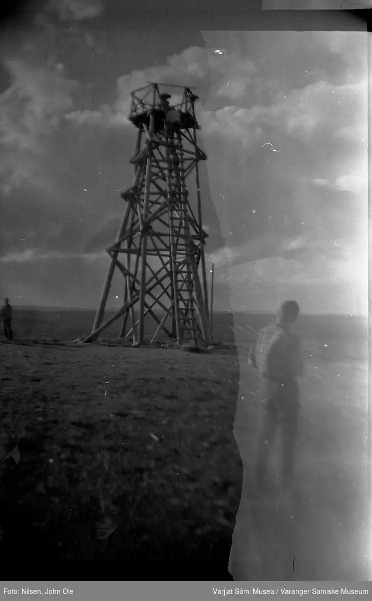 Muligens et utkikkstårn. En mann står med ryggen til i forgrunnen og en annen mann klatrer opp i tårnet. Ukjent sted 9. juli 1966.