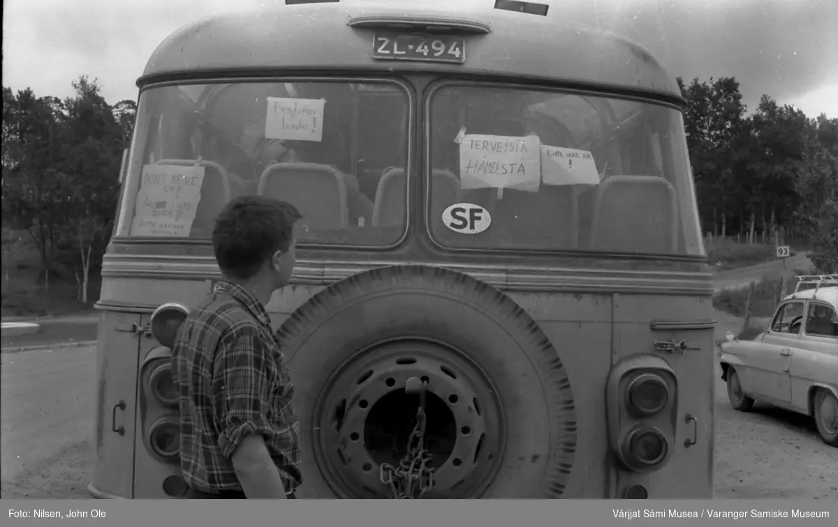 Oleif Nilsen Elen kikker på plakater i bakvinduet på en finsk buss som står på en parkeringsplass. Ukjent sted høst 1966.