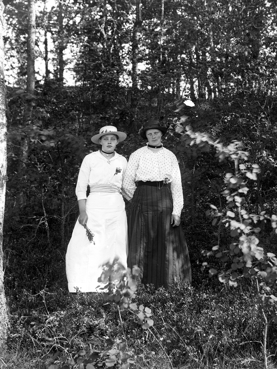 Systrarna Agnes (till höger) och Ellen Sikén fotograferade i en skogsbacke i Karl Gustav. Agnes har en ljus dräkt och ljus hatt. Ellen har mörk kjol och en mörk hatt.