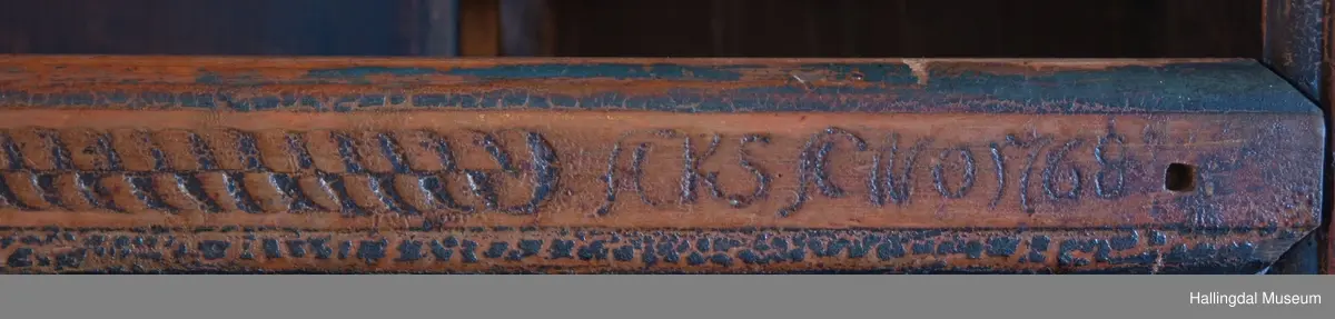 Rester etter rød og blå maling.  Gesims, skråner ned bakover. Profilert. 6 sirkulære hull i arkitraven.  Mellom disse er malt med sorte bokstaver.  Gavlene har utskårne "hylleknekter".  Bakveggen består av fire vertikale planker.  Skjøtene er dekket med profilerte lister.  Bare to biter i nederste del til venstre er fremdeles på plass.  Nederste del er lukket skap.  Tredelt, midterste del er skapdør.  Platen har profillist.  Feltene har speilfyllinger.  Blåmalte.  Klosset marmorering.  Speilfyllingenes form er som en liggen H.  Har profillist.  Skapet er satt på en ny grønmalt plate.  På døren er malt hvite bokstaver.  Nederst på døren er skåret inn bokstaver, opp-ned.  To nøkkelhull m. jernbeslag på venstre side av døren.  Over døren til venstre, en dreibar trekloss, holder igjen.  Innvendig har døren merke etter stort jernbeslag på samme side som nøkkelhullene.  To lister skåret i treverket minner om lange jernbeslag,-smalner mot nøkkelhullene.  Akantusranker i rødt, blått og hvitt, meget avslitt.  En hylle i skapet. Mangler bakvegg.  Karmen har på venstre side spor etter to jernhengsler.  Øverst på høyre kortside, mangler gesimsen en bit, og holdes til sideveggen med en jernkrampe.  Jernkrampe også på venstre side.  Skapplaten mangler list på høyre side.  Venstre side, fra gesimsen og ned har spor etter å ha stått inntil en vegg eller et annet møbel.