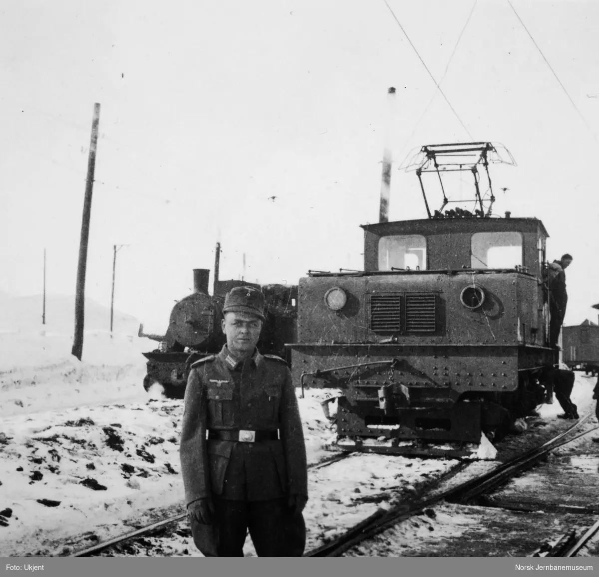 Tysk soldat foran et elektrisk lokomotiv på Sydvarangers jernbane, med et damplokomotiv i bakgrunnen