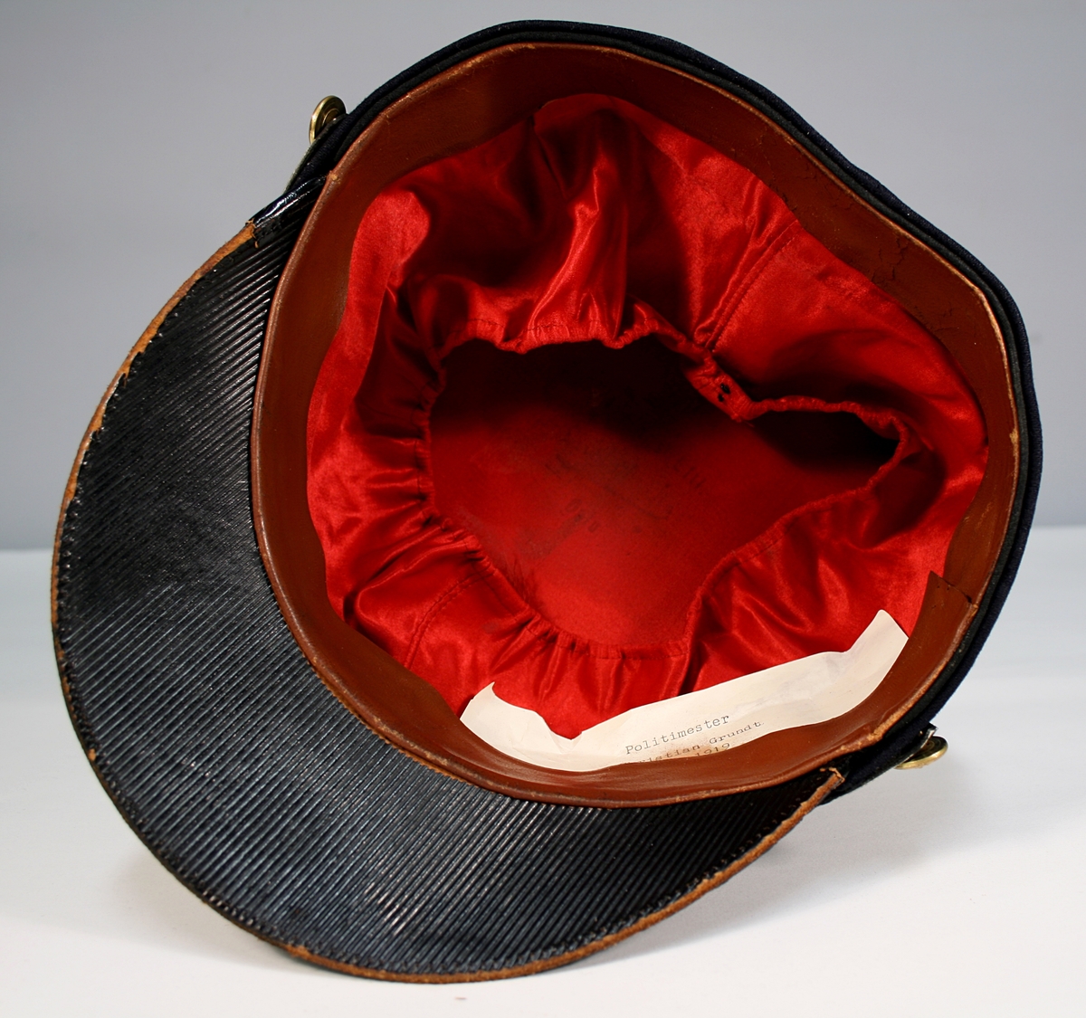 Sort kepilue med riksvåpen i metall omgitt av gullbrodert åpen ekebladskrans. Gullpompon. Smalt sort luebånd med gullbroderte ekeblad og ekenøtter. Reim og skygge isort lakk.