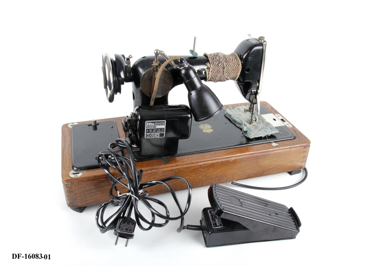Symaskin på tresokkel, med tilhørende lokk med lås og håndtak. Symaskinen er utstyrt med lampe og  elektrisk motor, festet på baksiden med ledning og pedal.