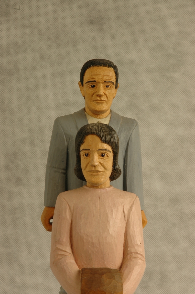 Skåret og delvis malt trefigur på sokkel. Figuren består av en mann og en kvinne. Begge personfigurene er løst nedfelt i sokkelen.