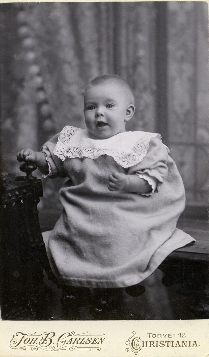 Portrett av en liten baby. Babyen er iført hvit kjole med en hvit krage.