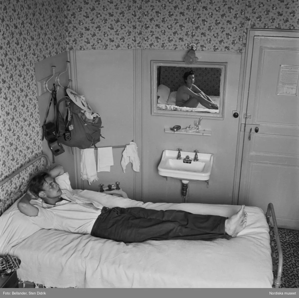 Hotellrum. Fotografen själv ligger i sängen och i spegeln syns hans hustru Birgitta. Ryggsäck och kamera hänger på krokar på väggen.