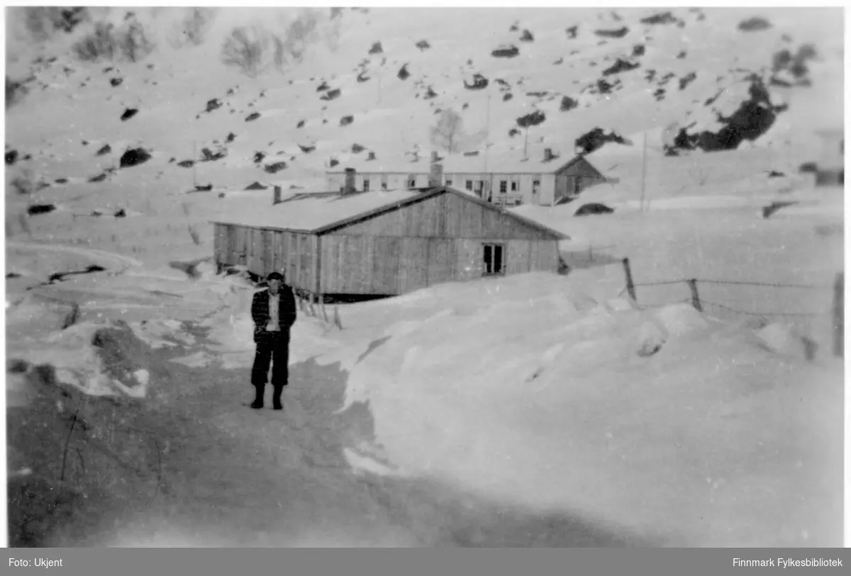 Brakkerbygninger i Øksfjord etter krigen, bildet tatt ca. 1946. En mann står foran Belsenbrakka som ligger nærmest stranda. Bak den ligger Sykestuebrakka. Ved siden av den nærmeste brakken er det blitt satt opp et gjerde. Man kan se masse snø på bakken og fjell i bakgrunnen.