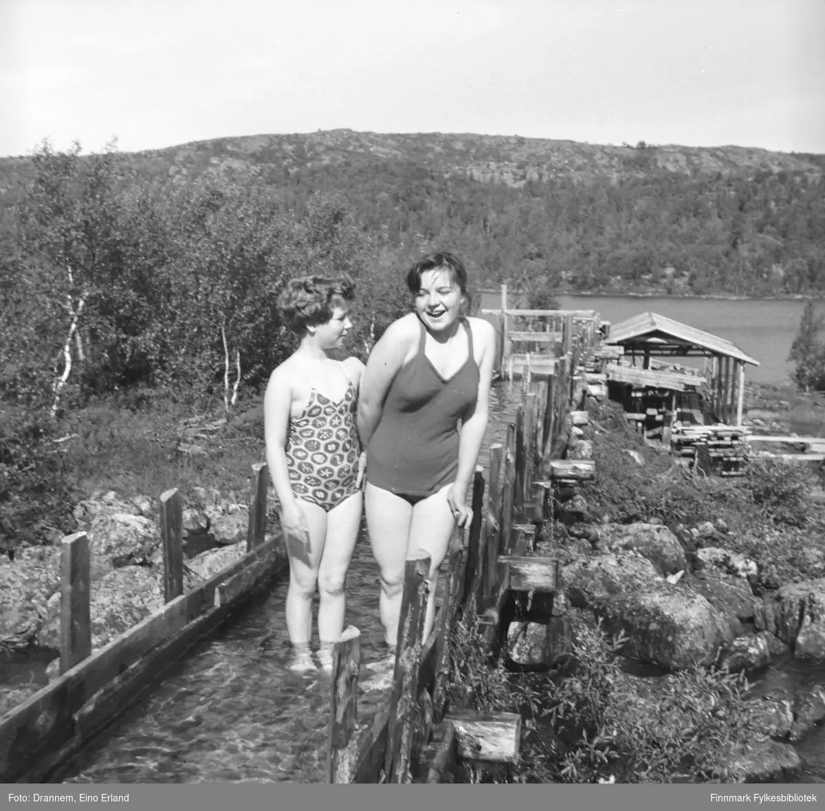 To jenter iført badedrakter, fre avenstre Turid Karikoski (Drannem) og Maija Hoikka (født Gerasimoff), står i en vannrenne som går til noe som ser ut som et lite sagbruk. 