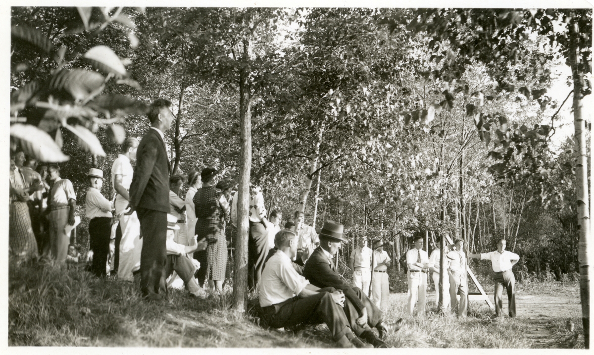 Bildet viser kvinner og menn som tilhørere/publikum. De står under noen trær i en park (?) Bildet er tatt i forbindelse med et stevne i Amerika i 1934. Alle er kledd i pent sommertøy. Olaus Islandsmoen sitter foran med hatt.