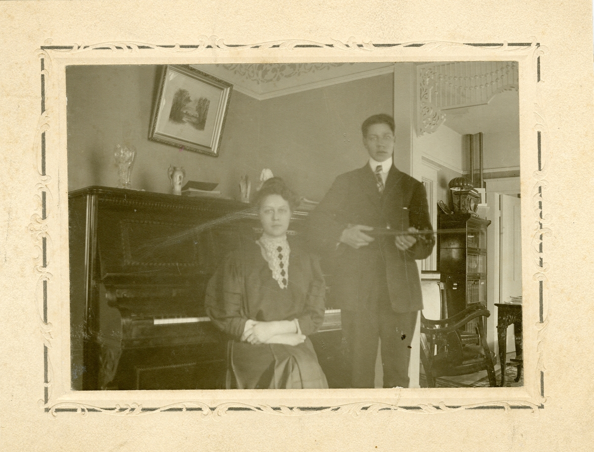 Bildet viser en ung kvinne sittende ved et piano og en ung mann stående ved siden av. Mannen holder en fele/fiolin under armen. Dette er norskamerikanere og foto er tatt i USA.