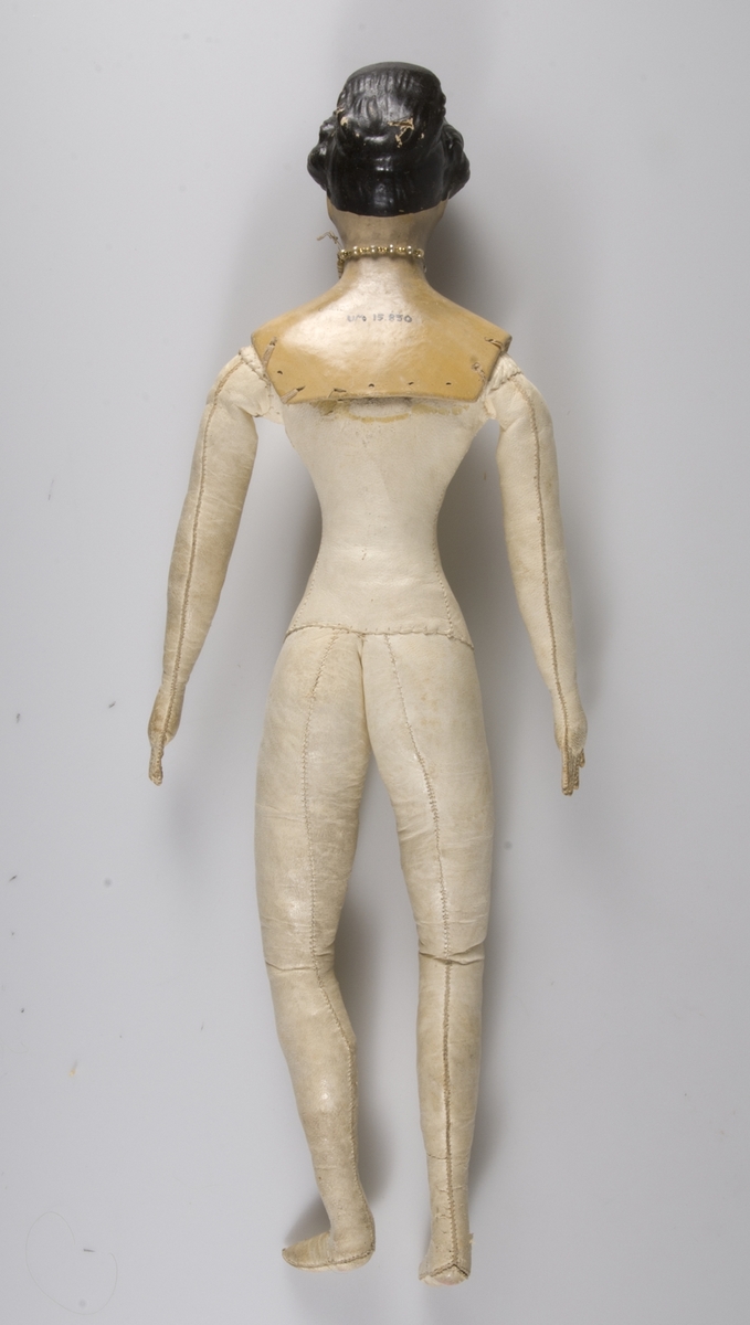 Docka. Huvud av papier-maché med svart målat hår och blå ögon. Kroppen stoppad, klädd med vitt glacéskinn. Iklädd klänning UM15856 och underkläder UM15852-15853.