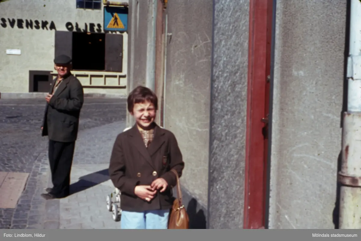 Lisbeth Sjöberg, dotter till Märta Sjöberg, fotograferad på trottoaren utanför Rasmussons skoaffär med adress Kvarnbygatan 4 vid Gamla Torget i Mölndal, ca 1961-1962. Längre bort på gatan står en man vid ett övergångsställe. I bakgrunden ses del av gaveln till huset Kvarnbygatan 6, byggnad tillhörande SOAB.

För mer information om bilden se under tilläggsinformation.