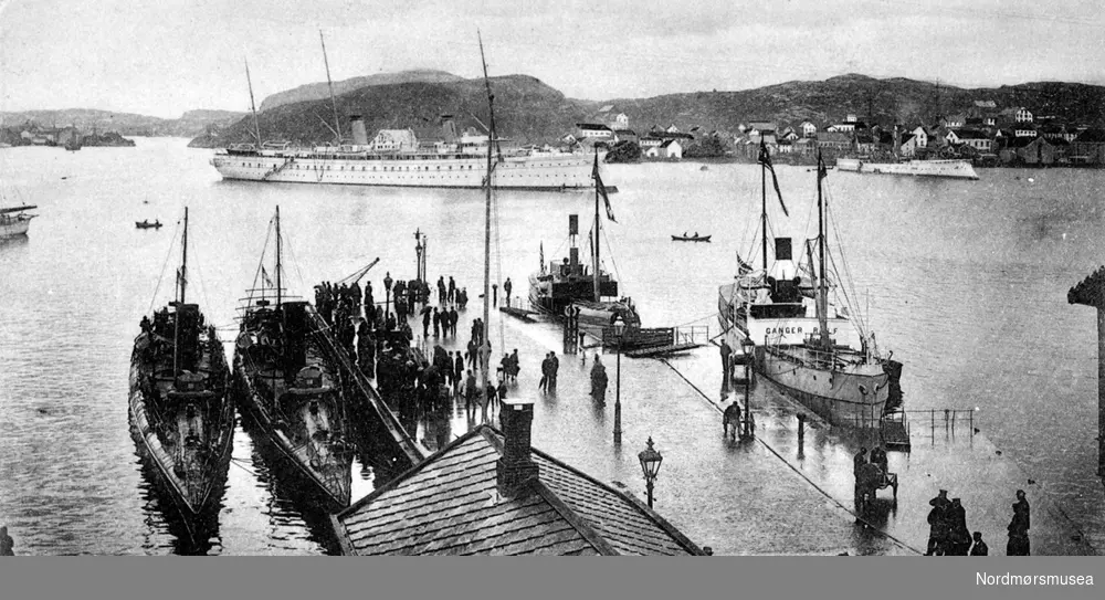 postkort "Den tyske Keisers Besøg i Kristiansund". Her ser vi fra Kirkelandet Allmenning, hvor vi ser to kanonbåter liggende fortøyd. I bakgrunnen ser vi Keiser Wilhelm II's båt, med et større krigsskip til venstre(?). Til høyre for Allmenningen ligger Ganger Rolf og en til av rutebåtene. - I bakgrunnen ses, fra venstre, Nordholmen i Nordsundet og Nordlandet.  Fotograf er Johan K. Engvig, og bildet er datert omlag 1904. Fra Nordmøre Museums fotosamlinger.