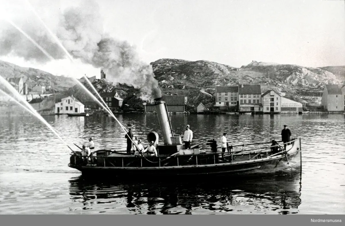 Brannsprøyta Slukk 1 som ble anskaffet i 1894 prøves ut i Vågen. Brannsprøyta kostet da omlag 40.000 kroner. I bakgrunnen ser en bebyggelsen på Gomalandet, med blant annet Woldbrygga og Likvoren. Ellers så ser vi på båten blandt annet følgende personer fra venstre: Havneformann P. Bugge, fører F. Knudsen, Brannassisten A. Sakshaug, K. Røsandhaug(?), Ukjent, Ukjent, Maskinist F. Wilf(?), ukjent, Byigeniør Bäckstrøm. Fra Nordmøre Museums fotosamlinger.