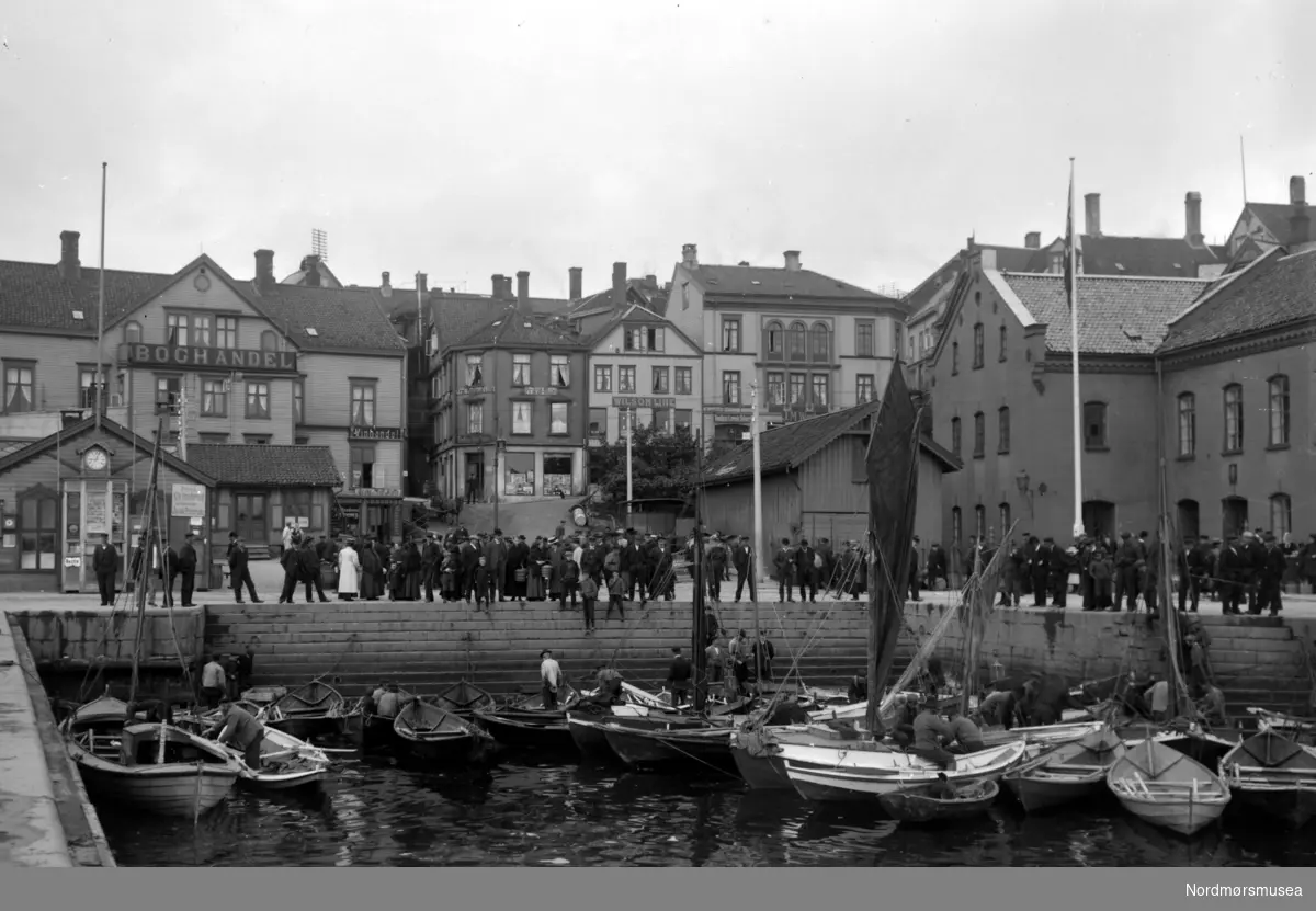 Et bilde fra Fisketrappa og Fisketorget på Kirkelandet i Kristiansund. I Fisketrappa ligger robåtene som "sild i tønne". Kaien er også fylt med mennesker, med forskjellige forettninger langs kaien. Vi ser i bakgrunnen blant annet Heinrich Loennechens bokhandel (til venstre) samt Tollbua (til høyre). Arnesens skipshandel foran. Nordmøre Museums fotosamling