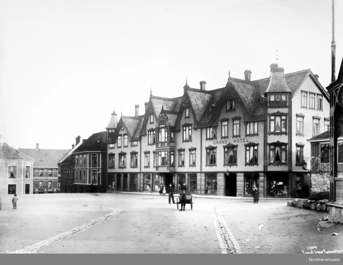 Det første Grand Hotell

Etter en alvorlig økonomisk krise i 1880 årene var byen nå atter preget av fremgang og optimisme. Byen hadde på denne tid en befolkning på 11 til 12000 innbyggere. Det var nå behov for et nytt standmessig hotell for reiselivsnæringen, foretningsfolk samt byens egen befolkning.

Gustav W. Dahl som var født i Trondheim i 1863, der han i første halvpart av 1890 årene hadde arbeidet som hotelltjener blant annet ved Grand Hotell cafe. At han ville prøve seg på hotelldrift i Kristiansund skyldes nok mye hans kone som var fra Kristiansund, og gjennom henne var blitt kjent med byen. 

Dette ledet til at i første halvpart av 1890-årene ble det oppført et boligkompleks på tomten til den gamle Prestegården, og som hadde fasade mot Torvet og Bernstorffstedet. Det var Stadskonduktør Hagbart Brinchmann som var byggherre og eier av bygget, hvor Grand Hotell etablerte seg i.

Mai 1895 kunne man lese i en annonse i to av byens aviser at byens Grand Hotell åpner dørene 1. juni 1895, og annonsen var signert hotellets eier Gustav W. Dahl. 

Hotellet hadde 30 værelser, og foruten gjesterom hadde det udpakningsværelser, en elegant Salon, Læseværelse og Spisesal.

Driften av hotellet ser ut til å ha godt bra, for i 1897 etablerte Gustav W. Dahl seg som restauratør på Øvre Bakklandet i Trondheim. Dette kjøpet ble fulgt opp av et nytt kjøp våren 1900, da han også kjøpte Trondheims Grand Hotell.

Ettersom Gustav W. Dahl nå hadde satset så hardt i Trondheim ble det behov for en ny innehaver for Grand Hotell i Kristiansund. 20. november 1900 kunne man da også lese i avisen i Kristiansund at nye lederen for hotellet nå var Nils Nilsen.

Nils Nilsen var født i Sand i Ryfylke i 1869. Han hadde vært reisefører og tolk for den britiske godseier og vinkongen Fleetwood Sandeman og gjennom hans reiser gjennom flere europeiske land, hadde fått et inngående kjennskap til hotelldrift. På det tidspunkt han tok over driften av hotellet var han faktor ved Sandemans gård Gulla i Surnadal. Trolig fikk Nils Nilsen også økonomisk støtte fra sin arbeidsgiver til å kjøpe hotellet.

En av forandringene Nils Nilsen gjorde ved hotellet var å installere hustelefon på alle rom.

En av de større begivenhetene hotellet opplevde var å være vertskap for kongefamilien på dens kroningsferd den 18. juni 1906. Nils Nilsen sørget først for en bedre borgermiddag i gymnastikksalen og i andre rom ved Enggatens skole. Hans Majestet Kong Haakon VII overnattet senere på hotellet, mens Hennes Majestet Dronning Maud overnattet på  kongeskipet Heimdal for å få mer ro neste morgen. Morgenen etter ble det arrangert en avskjedsfrokost for kongen, der medlemmer av formannskapet og embetsmenn med fruer deltok. 

Så i 1899 døde Hagbart Brinchmann som eide bygningen der Grand Hotell holdt til, og hans enke, Laura Susanne Brinchmann (født Øwre) solgte da bygningen til Nils Nilsen våren 1907. Nils Nilsen var nå blitt eier av både hotellet og bygningen.

Men så den 8. november 1907 brøt det ut brann i hotellets vedbu. Brannen ble oppdaget av nattevakten, og alarm ble slått. Mer enn 300 mann kom raskt til stede, samt 5 fra sjødampsprøyta, men det ble snart klart at det store trekomplekset ikke kunne reddes. To mennesker mistet livet i brannen. Disse var søstrene Anna og Augusta Sørensen som drev motehandel i første etasje av hotellet. Den flotte trebygningen i utført sveitserstil var nå blitt slukt av flammene.

Ikke lenge etter besluttet Nils Nilsen å bygge et nytt hotell på den samme tomten. Arkitekten ble Kristen Tobias Rivertz, og det nye hotellet som ble oppført i jugendstil, stod så ferdig januar 1909.

Bildet er publisert i 1902, noe som trolig også er fotoår. Fra Nordmøre Museums fotosamlinger.

Referanse: En opplevelsens festlabyrint av Egil Husby, side 7 - 16). Nordmøre Museums fotosamling.