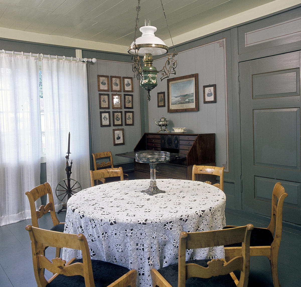 Interiør; rundt bord med hvit duk og stoler rundt i forgrunnen, husordel, mange fotografier på veggen og en oljelampe over bordet.