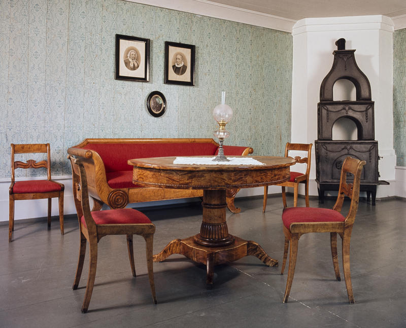 Interiør; brunt bord med stoler og sofa i samme treverk og med rødt trekk, to gamle fotografier på veggen og en etasjeovn i støpejern. (Foto/Photo)
